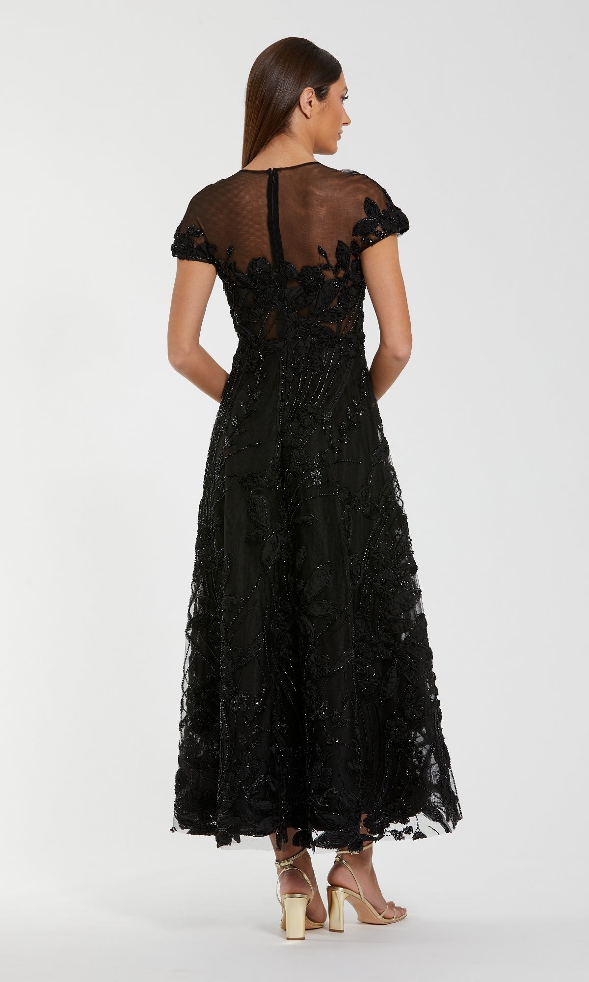 A woman faces away, wearing the Mac Duggal 69003 Cap-Sleeve Black Formal Dress with sheer fabric, floral accents, flared skirt, and gold heels.